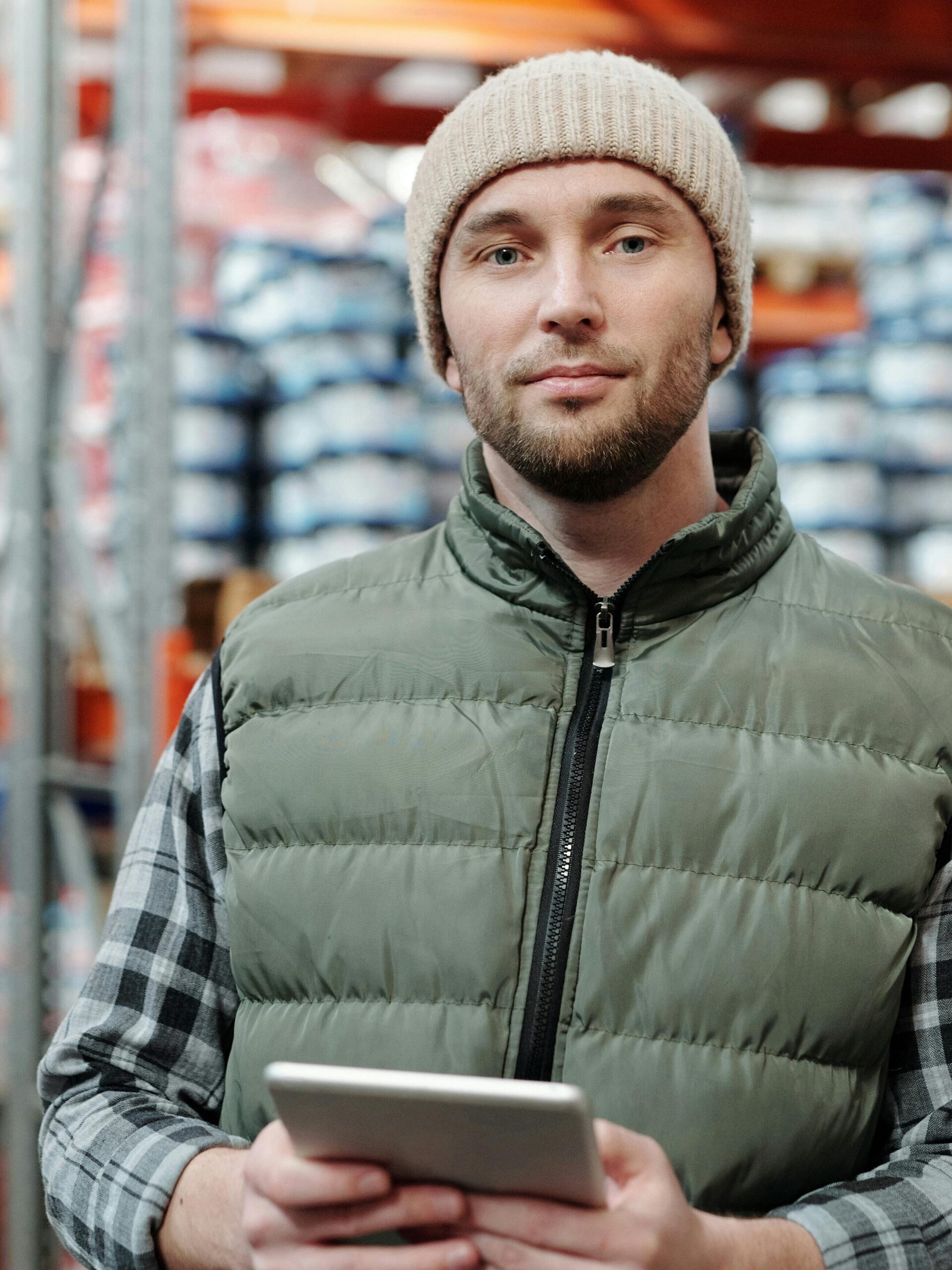 pexels-photo-4484072-4484072 A man wearing a beanie and vest holding a tablet in a warehouse setting.