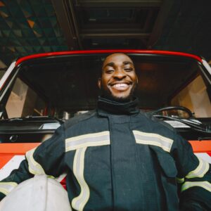 pexels-photo-7155779-7155779 Smiling firefighter in safety gear standing in front of a fire truck, conveying bravery and readiness.